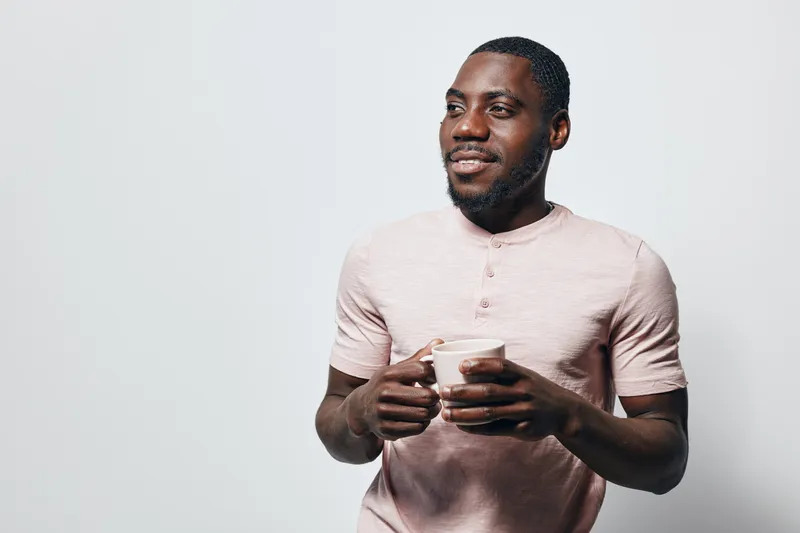 African American man with dark skin holding white cup, smiling and looking away. Casual clothing, short hair, standing against plain light background with relaxed and happy expression.