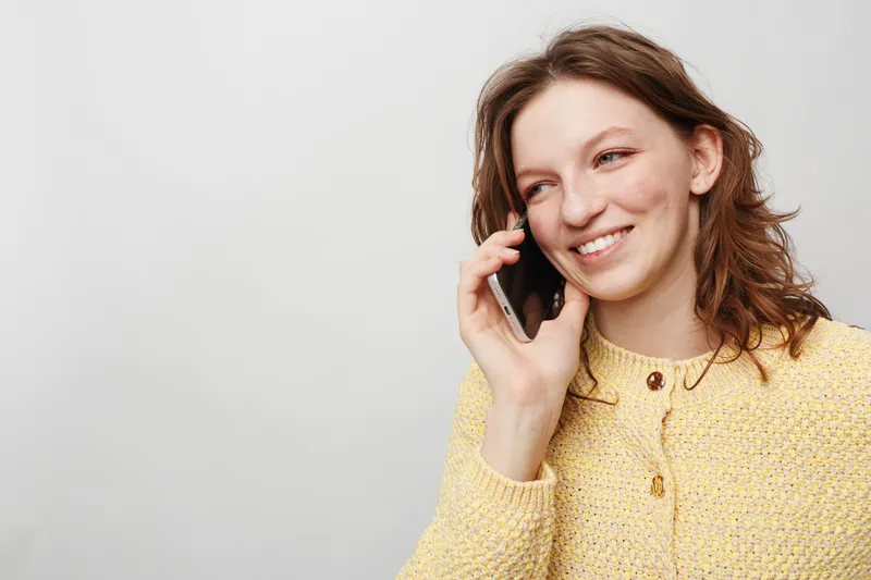 Smiling young woman with curly hair wearing yellow sweater talks on smartphone against light background. Concept of communication, technology, happiness, and casual lifestyle.