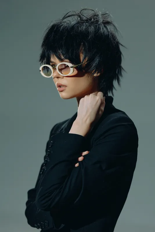 Fashionable woman with short hair and round glasses posing in dark clothing with a thoughtful expression.