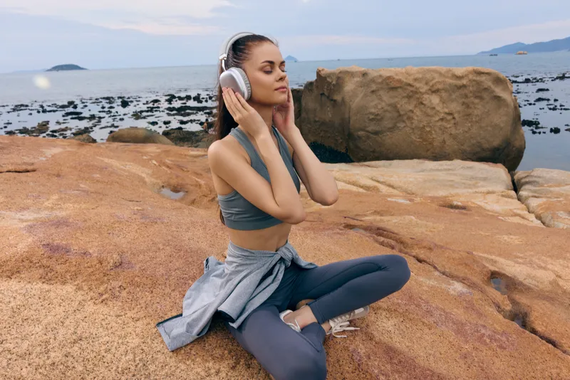 Young woman sitting on a rock by the sea, wearing sporty attire, headphones on, enjoying music with a serene expression, perfect for wellness and relaxation themes