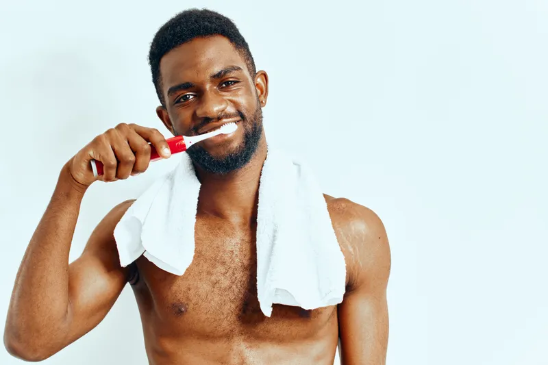Мужчина без рубашки сосредоточен на гигиене полости рта: он чистит зубы красной зубной щёткой в ванной, выполняя утренний ритуал