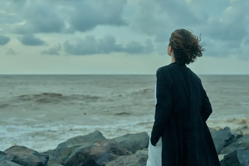 Woman in a dark coat stands on a rocky shore facing the sea, wind in her hair, contemplating the horizon and solitary water under cloudy skies. Soft.