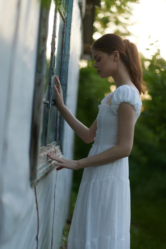 Young woman in a white dress contemplating by an old window in a serene outdoor setting with soft evening light