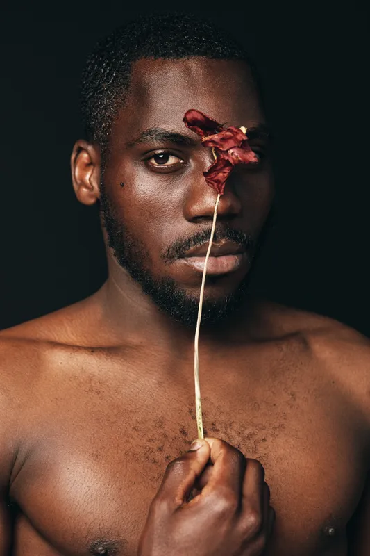 African man with dark skin holding dried flower in front of face in creative conceptual visual style. Portrait with bare chest and artistic expression on black background.