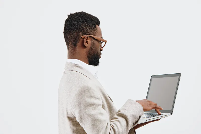 Professional young man with glasses using a silver laptop in a minimalistic studio setting, dressed in casual beige sweater. Focused on work or browsing, modern technology concept, white background.