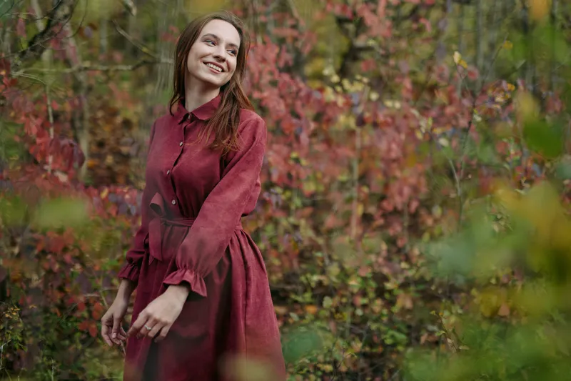 A candid portrait of a smiling woman in a burgundy dress amid autumn foliage, conveying authenticity through natural light, unposed posture, and genuine mood.