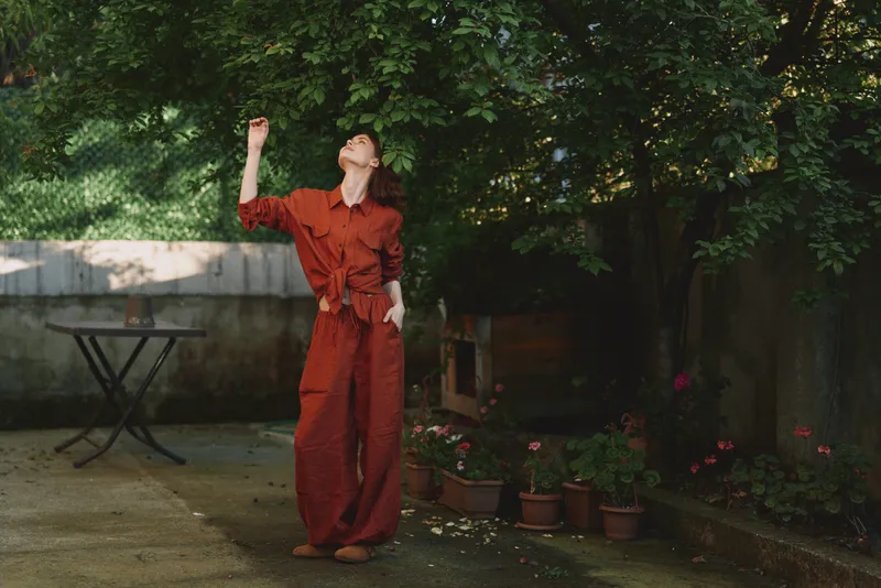 Smiling woman in a red jumpsuit stands in a sunlit garden, reaching upward with one arm. Outdoor scene highlights fashion, lifestyle, and relaxed self expression. Warm natural light