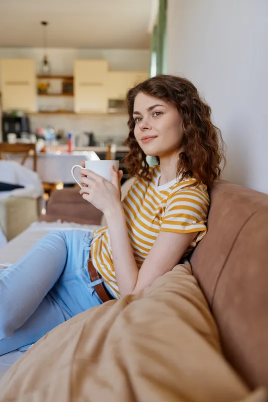 A woman relaxes on a sofa at home, sipping from a mug, wearing a striped tee and jeans. Her calm posture and soft lighting reflect comfort, routine, and personal downtime in a warm domestic setting.