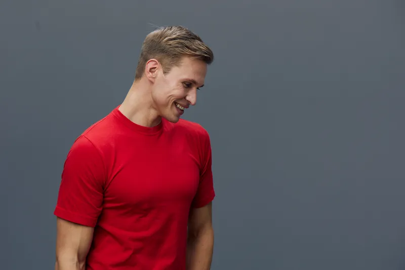smiling young caucasian man wear red t-shirt look down side isolated on gray background casual clothing digital detox concept