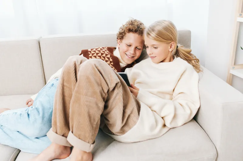 Authentic scene of children at home, siblings sharing a cozy sofa moment, exploring a tablet, embodying a playful school lifestyle and everyday curiosity, imagination.