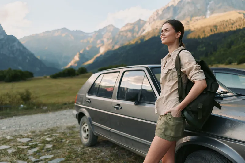 Young woman beside a parked SUV in a mountain valley, backpack on, ready for hiking, savoring a calm moment as towering peaks glow under golden light.