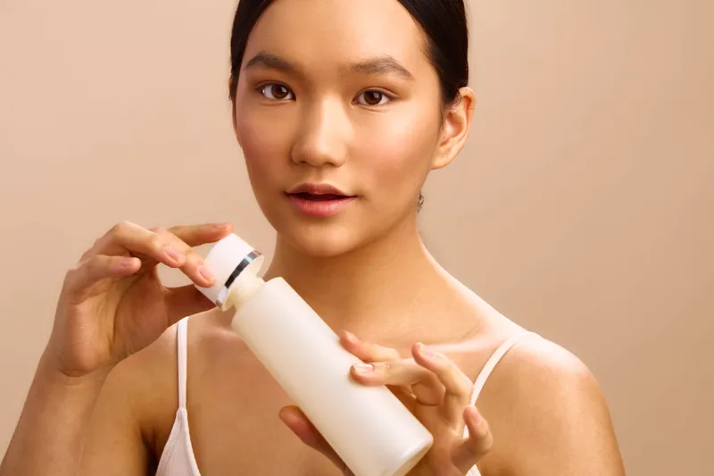 asian woman holds a skincare bottle in a modern setting, looking at the camera with a calm, confident expression, conveying self care, wellness, and a polished beauty routine.