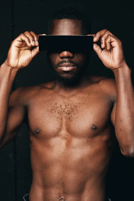 African man shirtless holding black rectangular object over eyes in creative conceptual visual style with dark background. Male model posing with bare chest and artistic expression.