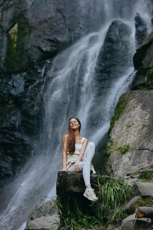 young woman sitting by waterfall in nature, wearing white clothes and sneakers, enjoying serene outdoor moment with greenery and rocks around, peaceful and relaxed atmosphere