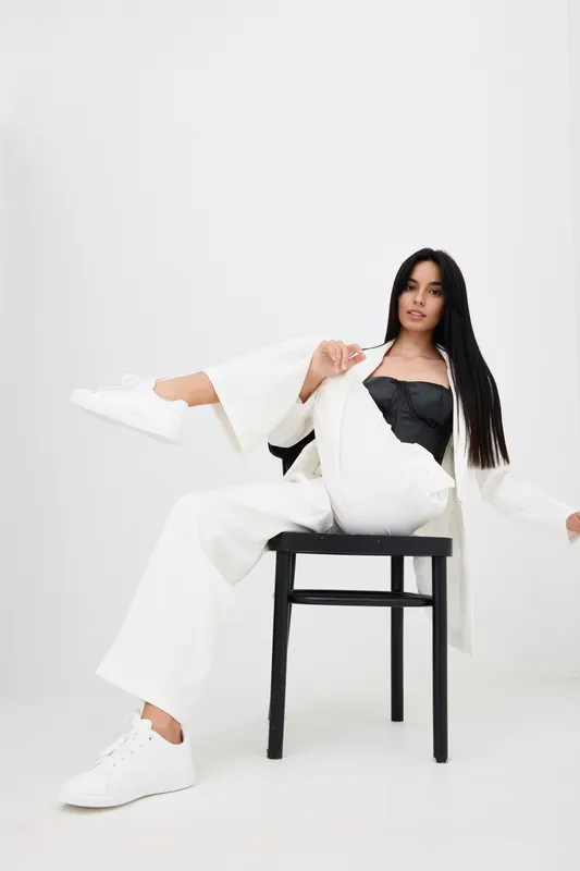 young woman in white wide-leg pants and jacket sitting on a black chair, wearing white sneakers and black top, casual style with relaxed confident pose in bright studio