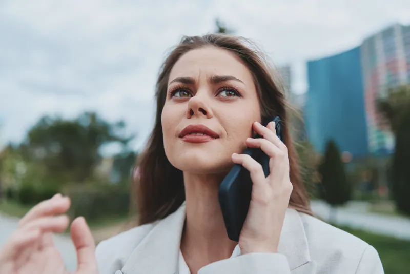 Confident young woman in white coat talking on smartphone outdoors with urban blurred background, business communication, emotion and technology concept