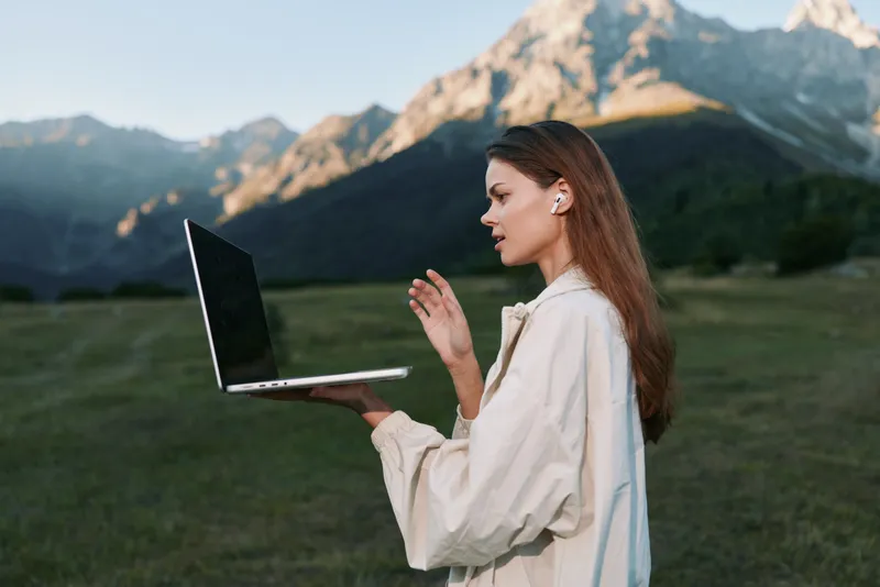 A thoughtful woman stands outdoors with a laptop in a rugged mountainous landscape. She focuses on the screen, projecting calm, confidence, and productive energy amid nature.