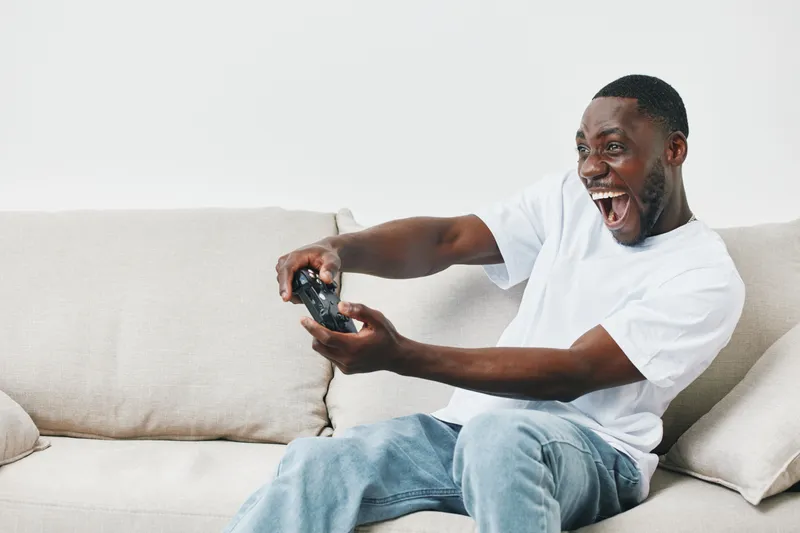 Happy young African American man enthusiastically playing video games on a couch, showcasing excitement and joy in a modern living room