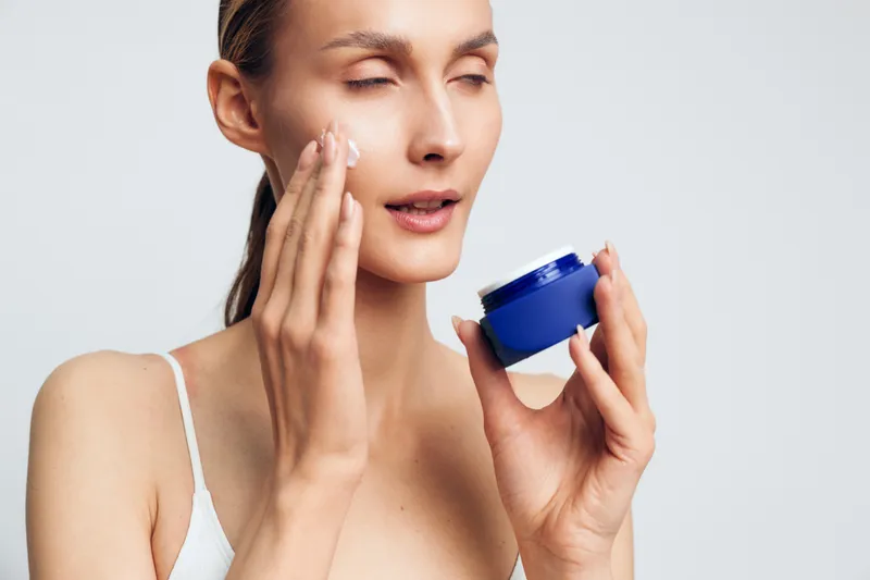 Woman skincare moisturizer cream facial hydration beauty applying to face close up, woman holds blue jar, soft studio white background, calm hydration and beauty routine