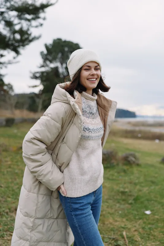 Woman outdoors wearing a long coat, knitted sweater and beanie, smiling in a park by the coast. Casual fashion, relaxed lifestyle portrait with nature backdrop