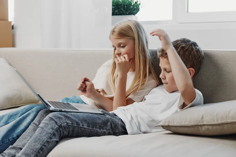 Two children relax on a beige sofa using a laptop, a boy excitedly gesturing while a girl watches thoughtfully. Bright living room setting with natural light from a window.