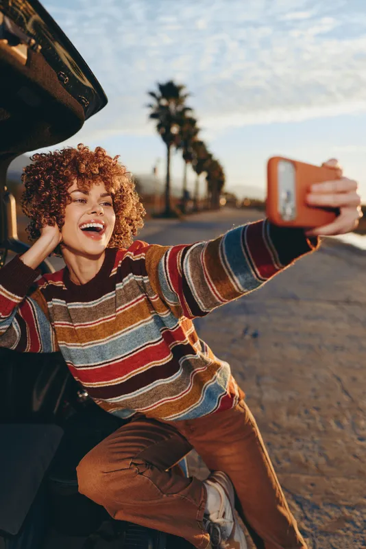 Woman selfie smartphone smiling curly hair outdoor street palm striped sweater, smiling woman holds smartphone for selfie on roadside, palm trees behind, travel lifestyle, golden hour