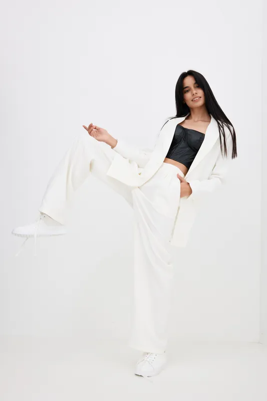 young woman in white suit posing confidently with raised leg, black top and white sneakers on plain white background, modern fashion style