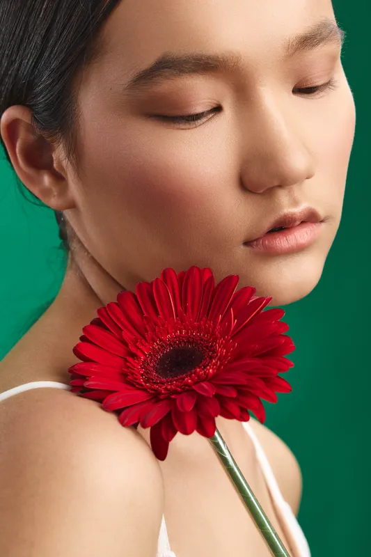 asian woman with delicate features stands against a rich green backdrop, holding a bold red flower near her shoulder. this serene beauty portrait emphasizes calm elegance and subtle artistry.