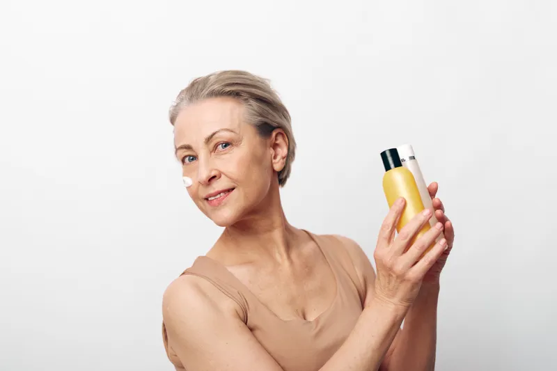 Smiling mature woman with short hair holding two bottles of skincare products, cream on face, isolated on white background. Concept of beauty, antiaging, wellness, and selfcare.
