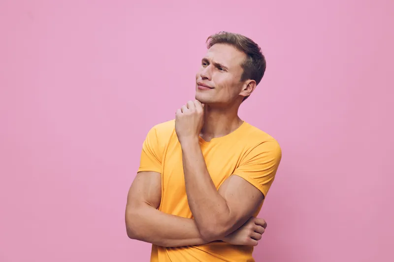Thoughtful mature man with short dark hair and a yellow t-shirt stands against a pink background, looking upwards with a pensive expression. Portrait, casual style, indoor studio shot, expressive, adu