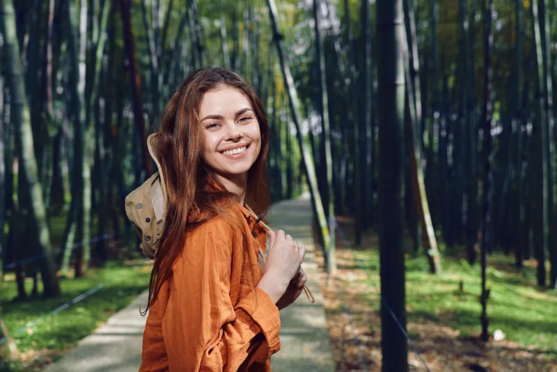 Woman smiling on a forest path holding a hat and backpack over shoulder, casual portrait in nature with sunlit trees and relaxed outdoor travel mood on a woodland trail.