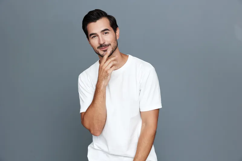 Handsome man in casual white t-shirt stands against gray background with thoughtful expression, touching chin. Portrait shows confidence and style.