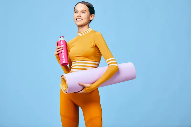 Fitness woman in an orange active suit with white striped sleeves stands against a blue studio backdrop, carrying a pink water bottle and a rolled purple yoga mat for a workout session.
