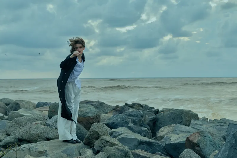 Windy coastal scene with a lone figure standing on jagged rocks beside the sea, wearing a light shirt and dark coat, facing the wind and spray.