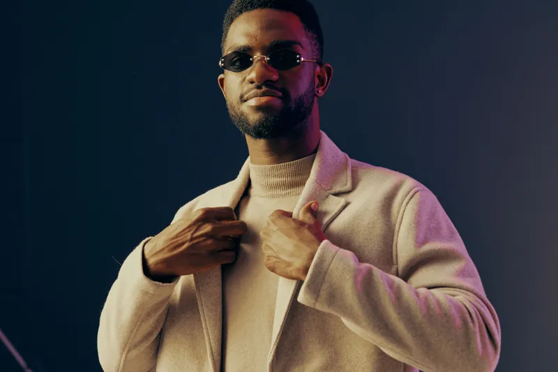A stylish young man of African descent wearing sunglasses and a light colored coat, exuding confidence against a dark background Fashion, elegance, and urban style