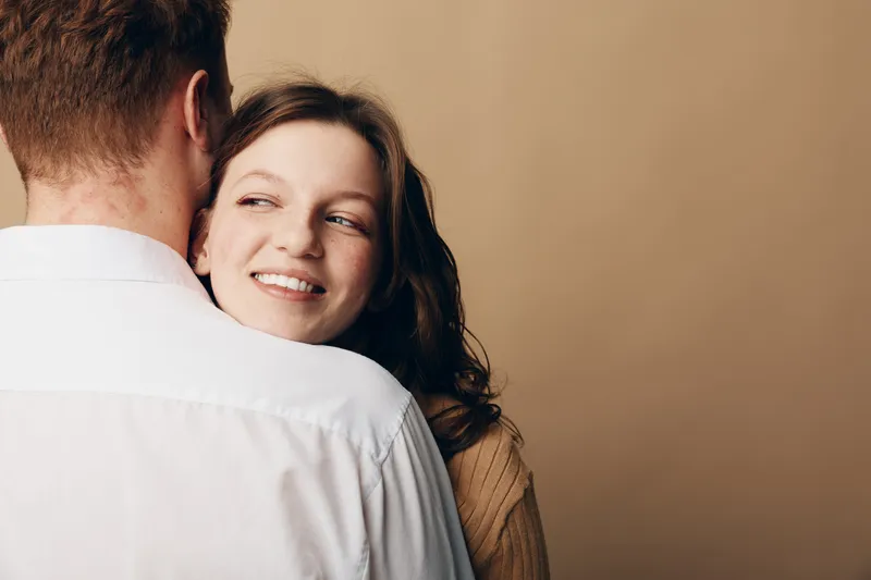 Smiling woman with long hair embraces man wearing white shirt, looking away. Warm beige background creates intimate and tender mood. Concept of love, relationship, and affection between couple.