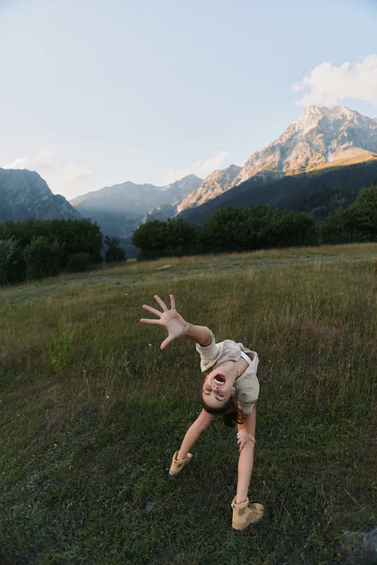 A young woman stretches into a playful backbend in a sunlit grassy meadow with towering mountains in the distance, capturing a spontaneous moment of carefree outdoor fun.