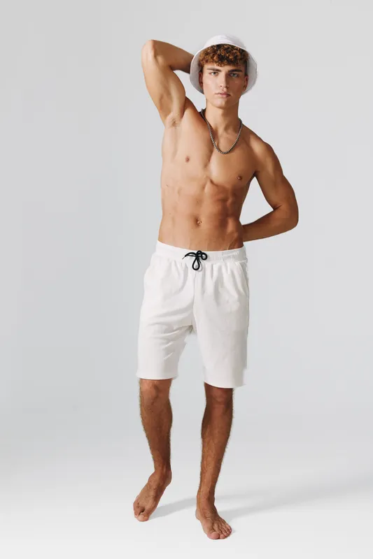 Young man with toned body wearing white shorts and bucket hat posing barefoot on gray background. Casual summer style with necklace and relaxed confident posture in studio lighting.