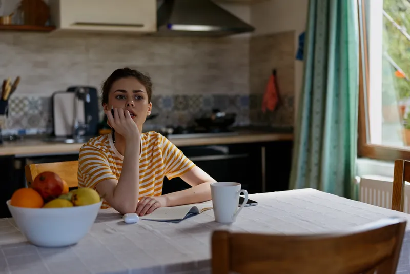 A thoughtful woman sits at a kitchen table, nibbling her nails while a notebook and a mug rest nearby. A fruit bowl adds color to a cozy home setting with soft daylight.