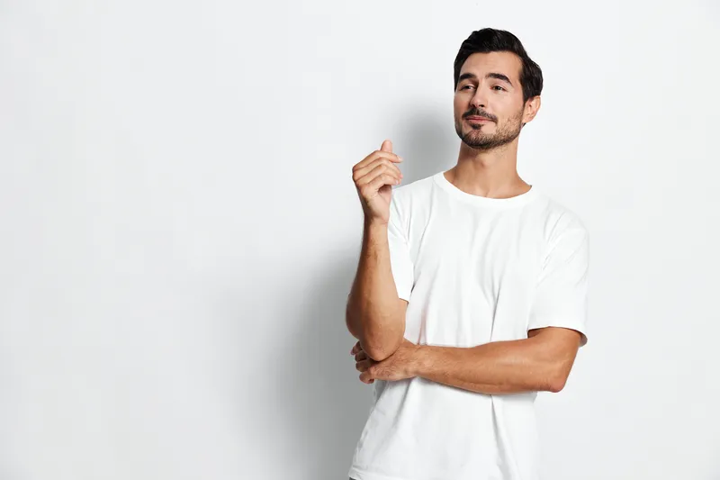 Full body confident young man in casual white t-shirt standing against plain solid background, smiling and gesturing with hand. Lifestyle concept.