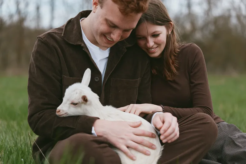 A young couple sitting on grass gently holds a white baby goat while sharing a tender moment in a natural outdoor setting during a casual lifestyle photoshoot.