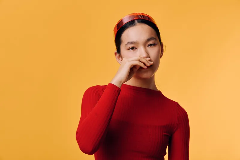 Young woman in red sweater and headband poses against orange background with hand near face looking thoughtful or contemplative in studio portrait with minimalistic style and soft lighting.