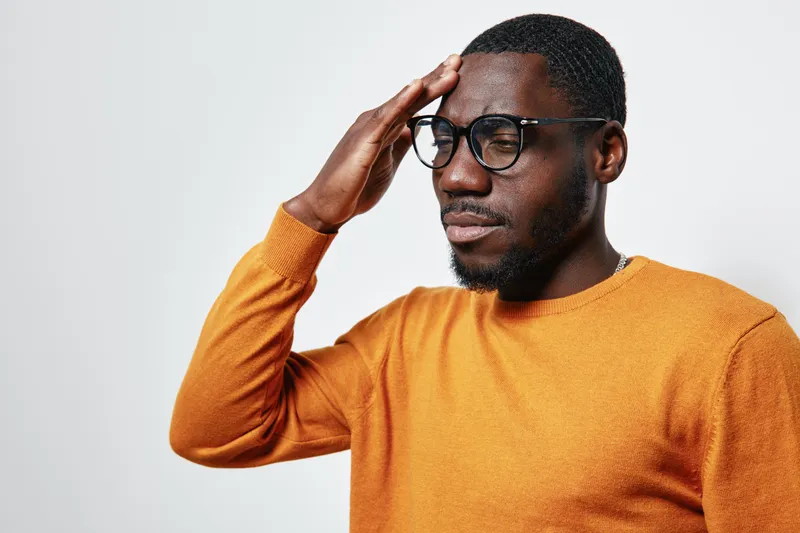 African American man with dark skin wearing glasses and orange sweater touching forehead with closed eyes, showing stress or headache on white background.