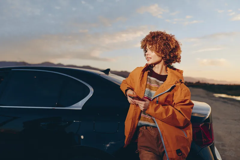 Woman lifestyle, car on road, sunset outdoors, curly hair woman holding bag, warm golden light, relaxed travel moment, scenic sky, orange jacket, casual outfit, evening road, calm mood