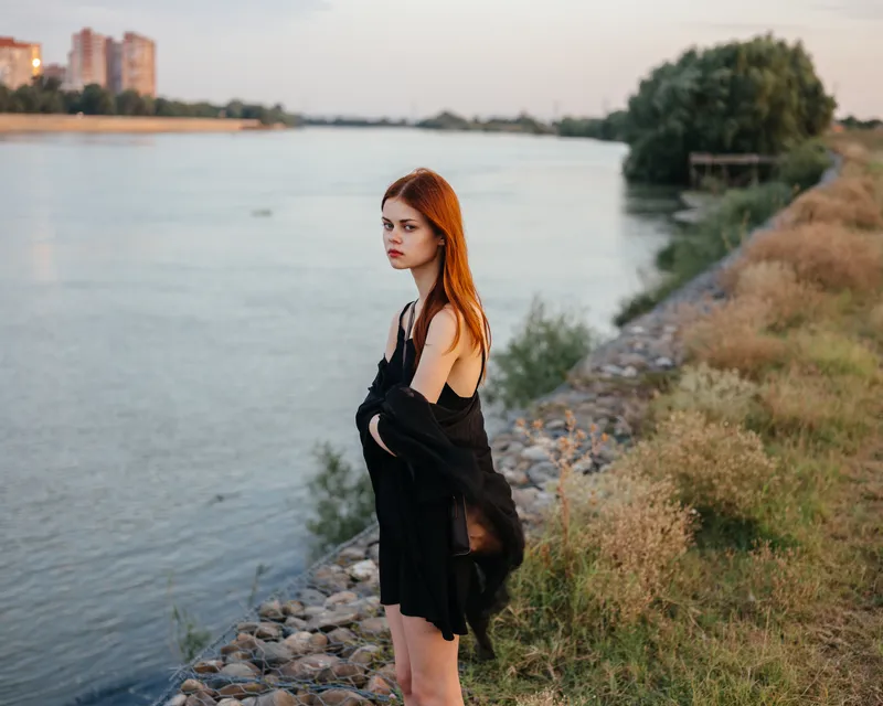 Рыжеволосая женщина в черном платье стоит у реки на природе