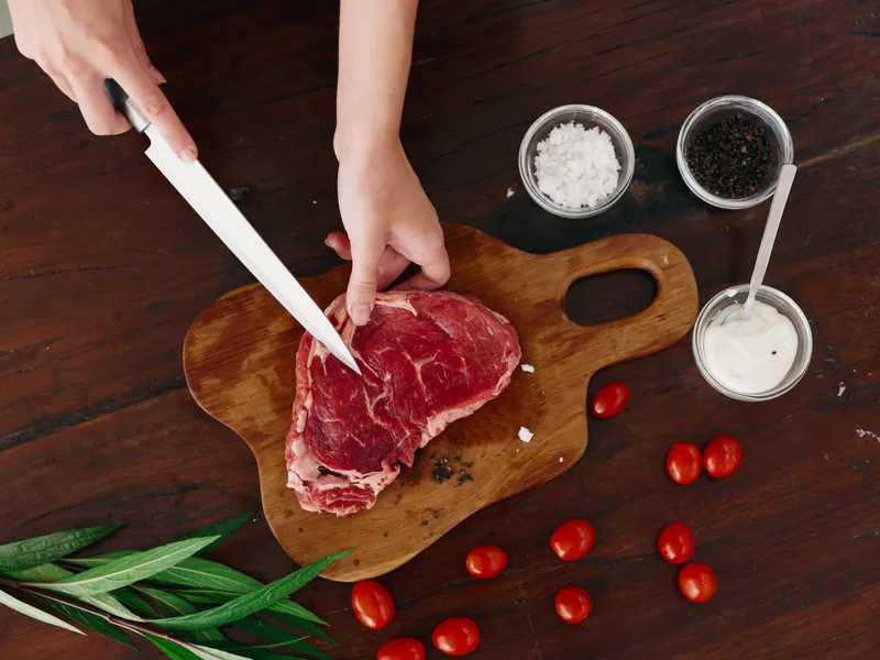 Женщина с ножом в руке нарезает свежее мясо стейка для жарки на кухне