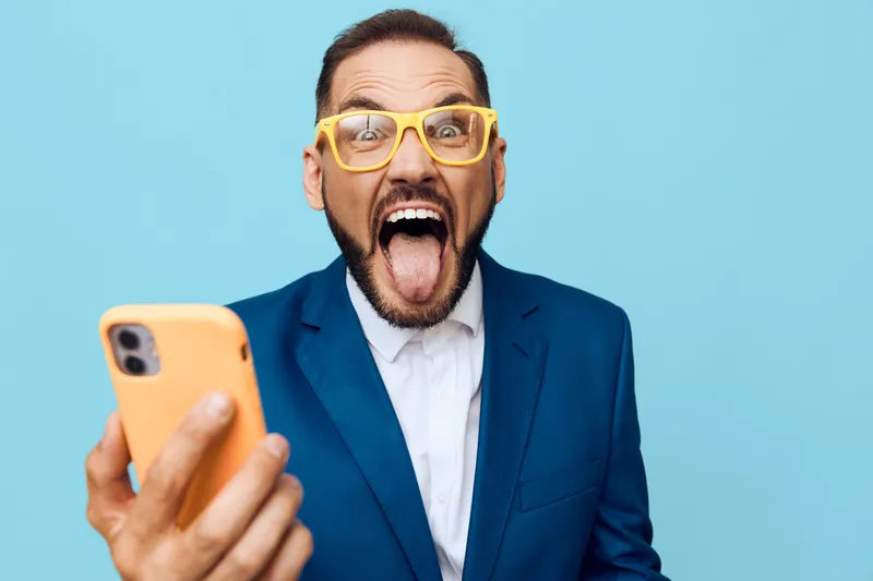 A business professional man on a solid blue background, exuding bold energy and excitement while looking at a mobile device with a wide open mouth, conveying strong professional presence.