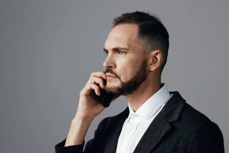 A focused business professional man on a solid colored background, portraying calm concentration and thoughtful mood while on a call, embodying confidence, strategy, and determination in a sleek