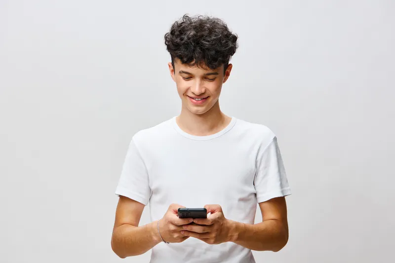 Улыбающийся молодой человек взаимодействует со своим смартфоном, одетый в простую белую футболку