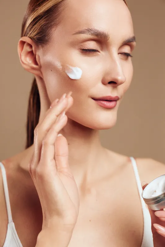 Young woman with closed eyes applying white face cream on cheek, holding container. Skincare and beauty concept with soft natural lighting on beige background.